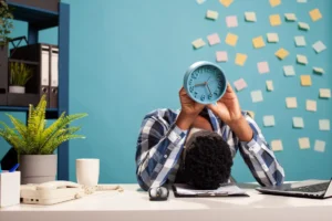 Funcionário com a cabeça sobre a mesa segurando um relógio, representando estresse e burnout no ambiente corporativo informal.