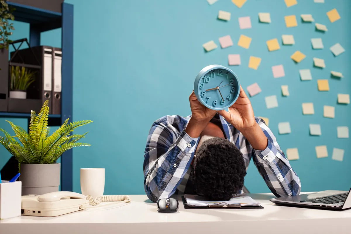 Funcionário com a cabeça sobre a mesa segurando um relógio, representando estresse e burnout no ambiente corporativo informal.