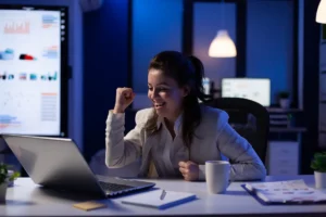 Mulher sorrindo e comemorando em frente ao notebook no escritório.