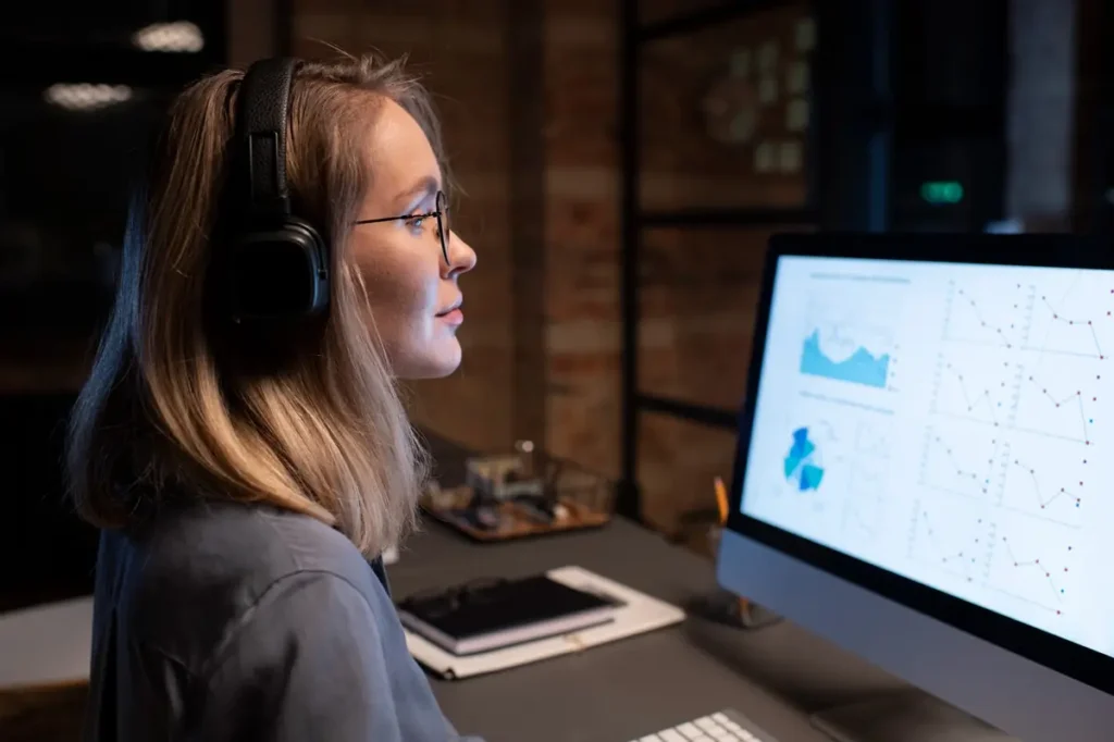 Mulher usando fones de ouvido enquanto analisa gráficos e indicadores em um computador, representando atividades de monitoramento de empresas.