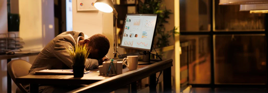 Homem cansado, de terno, com a cabeça sobre a mesa de trabalho enquanto observa gráficos em seu monitor, ilustrando um caso de burnout no ambiente corporativo.